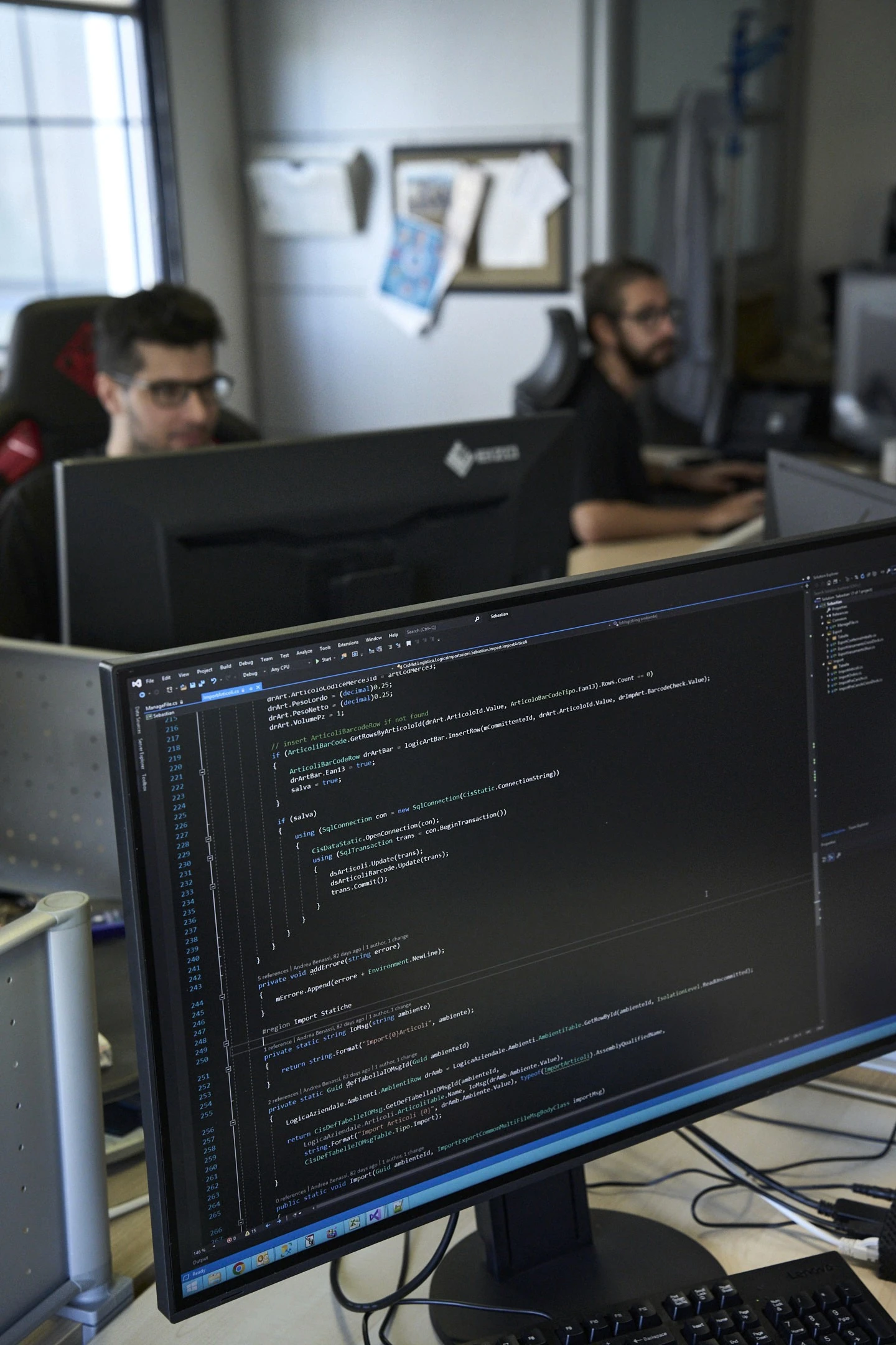 Developer working on warehouse and order management system logic at a desk, colleagues at desks in the background, in colour