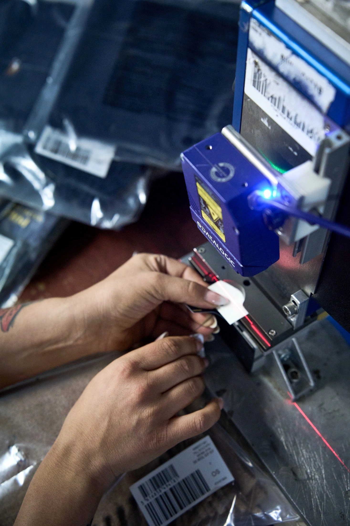 Hands position a garment tag under an industrial scanner, blue light and red laser beam, in colour