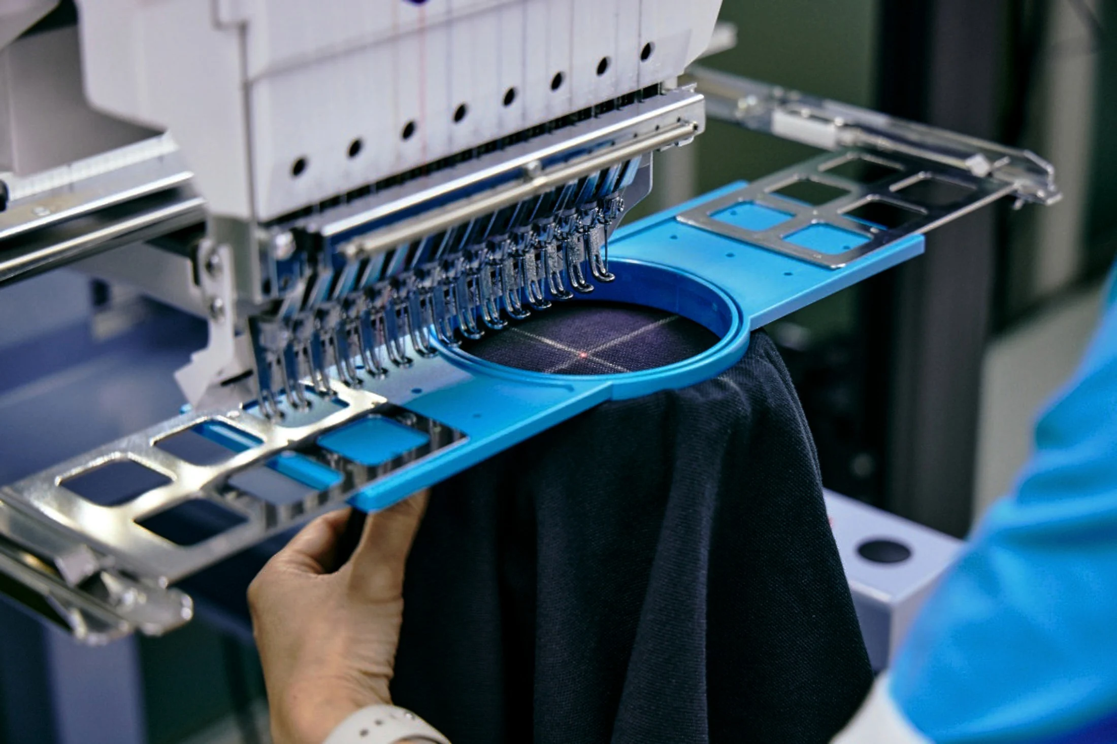 Hands positioning a dark garment under an embroidery machine head, close-up in colour